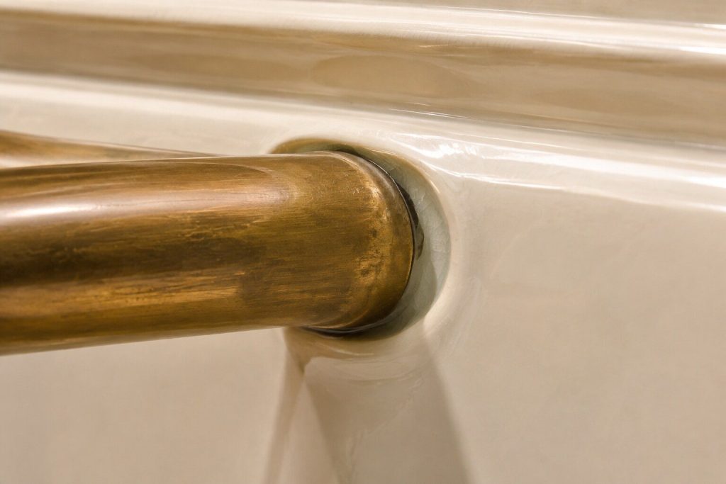 The joint where a heavy brass rail is let into the colored enamel front of a range, photographed very close in raking daylight so the hand-finished bevel and the depth of the glaze both show.