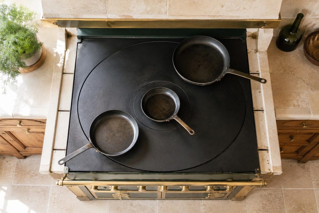 Three pans of different sizes standing at different distances from the center of a large cast-iron plate, seen from directly above in daylight — one over the middle, one halfway out, one at the cool rim.