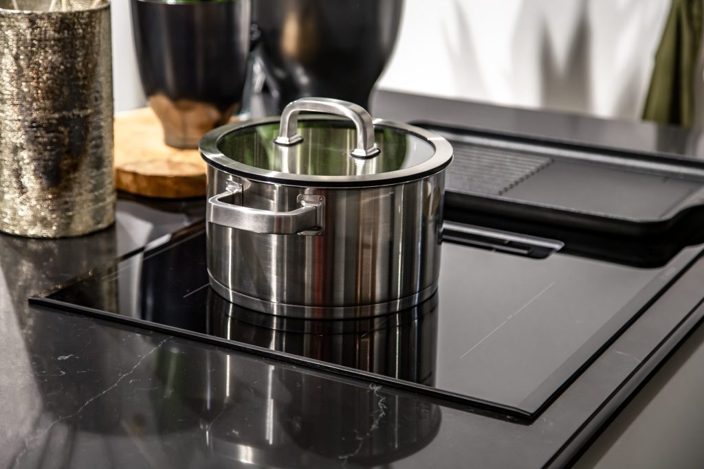 A steel pan standing on a black glass cooking surface set into a dark stone counter