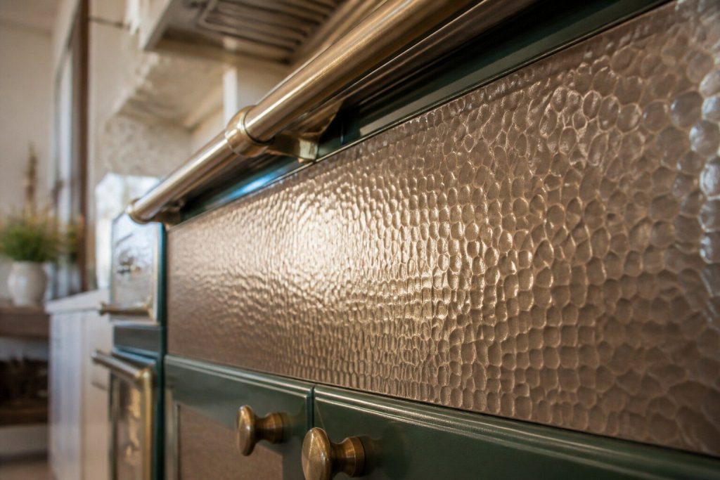 A hand-hammered metal panel on the front of a range seen very close, the dimpled surface scattering daylight into hundreds of small highlights across the whole frame.