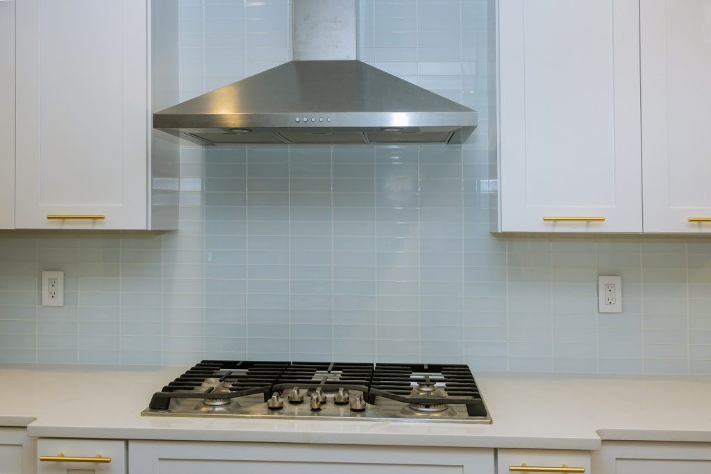 A stainless hood over a gas cooktop in a white tiled kitchen.