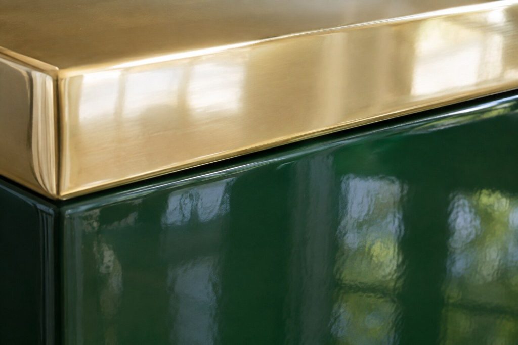 A very close raking view along the top corner of a hand-built range where a wide polished brass band meets a deep glossy colored enamel panel, the brass mirror-bright and the glaze holding a soft reflection of a window.
