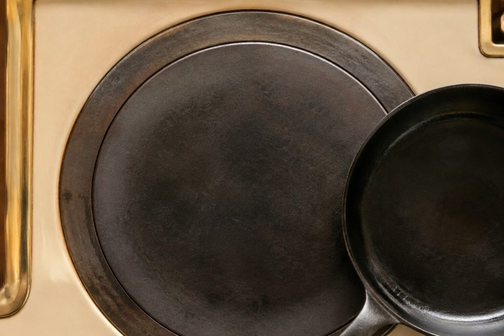 A thick round cast-iron cooking plate set into the top of a range, seen straight down from above in warm daylight, its center darker than its rim where the heat has been.