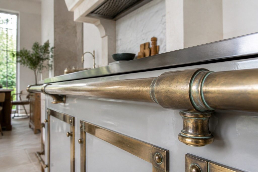 A close detail of an antique brass rail on the front of a range where the lacquer has gone cloudy and a faint green bloom has started in the crevices, photographed in bright daylight with the pale enamel out of focus behind.