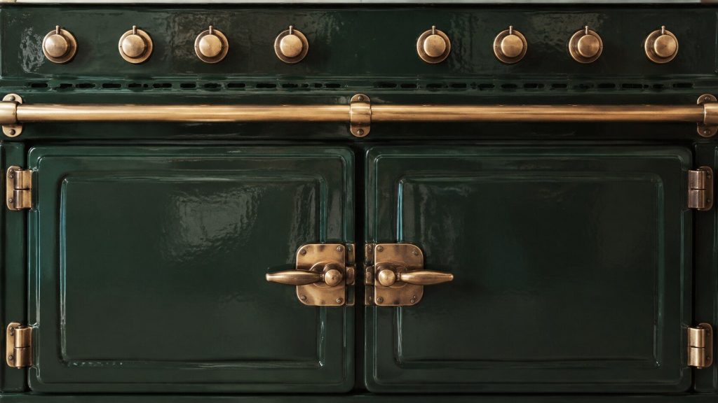 A tight square-on crop of the front of an enameled range, filling the frame edge to edge: two heavy oven doors below with their latch handles, a brass rail across the middle, deep colored glaze catching the window light.