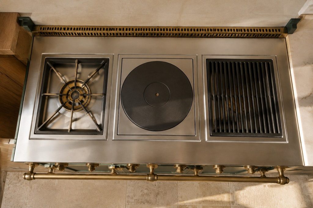 The top surface of a range seen from above, divided visibly into separate square modules — an open brass burner ring in one, a solid cast plate in another, a slatted grill in a third — each sitting in its own well in the steel.