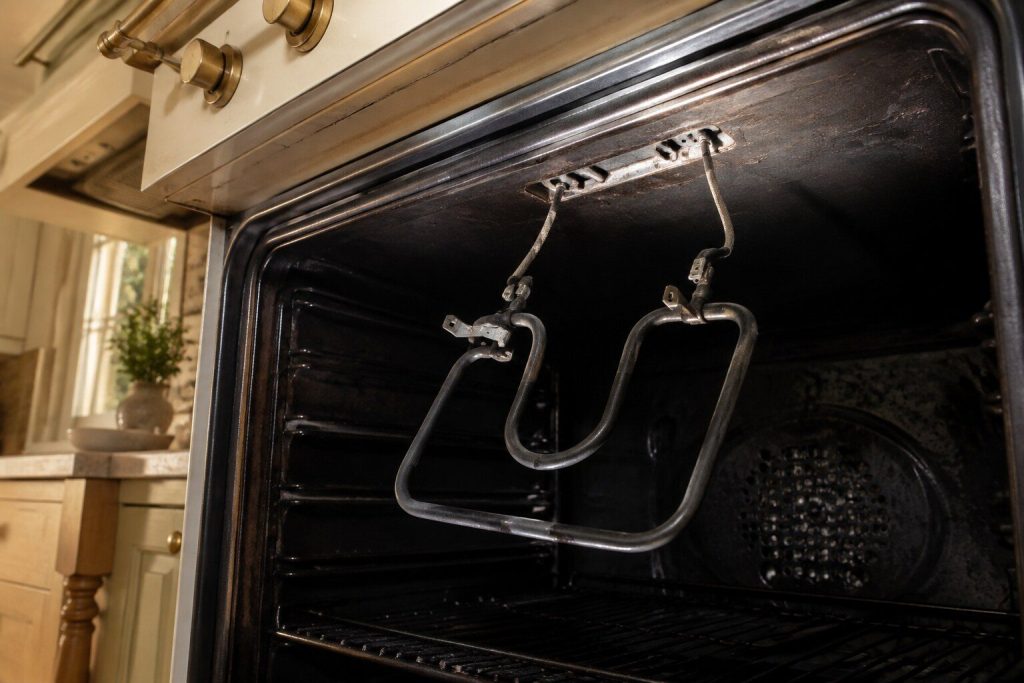 A curved electric broil element unbolted from the roof of an oven cavity and hanging down on its two leads, the empty mounting bracket above it.
