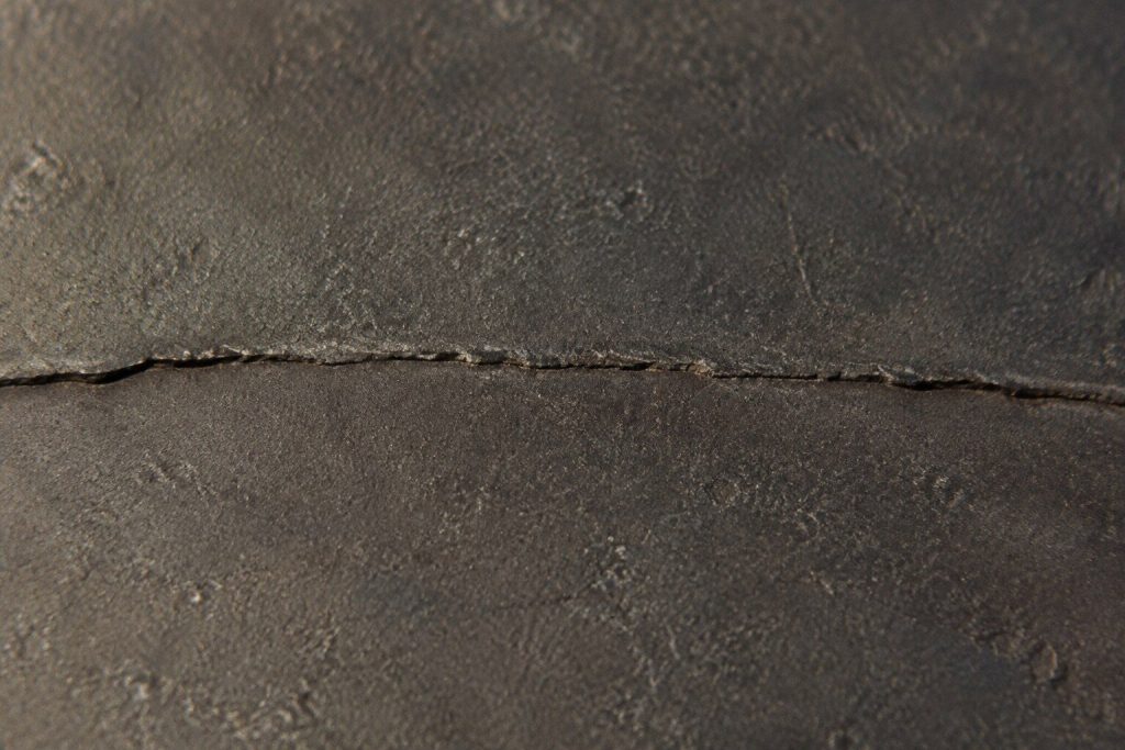 A fine crack running right across the surface of a heavy cast-iron cooking plate, photographed very close in raking daylight so the line and its lip catch the light.