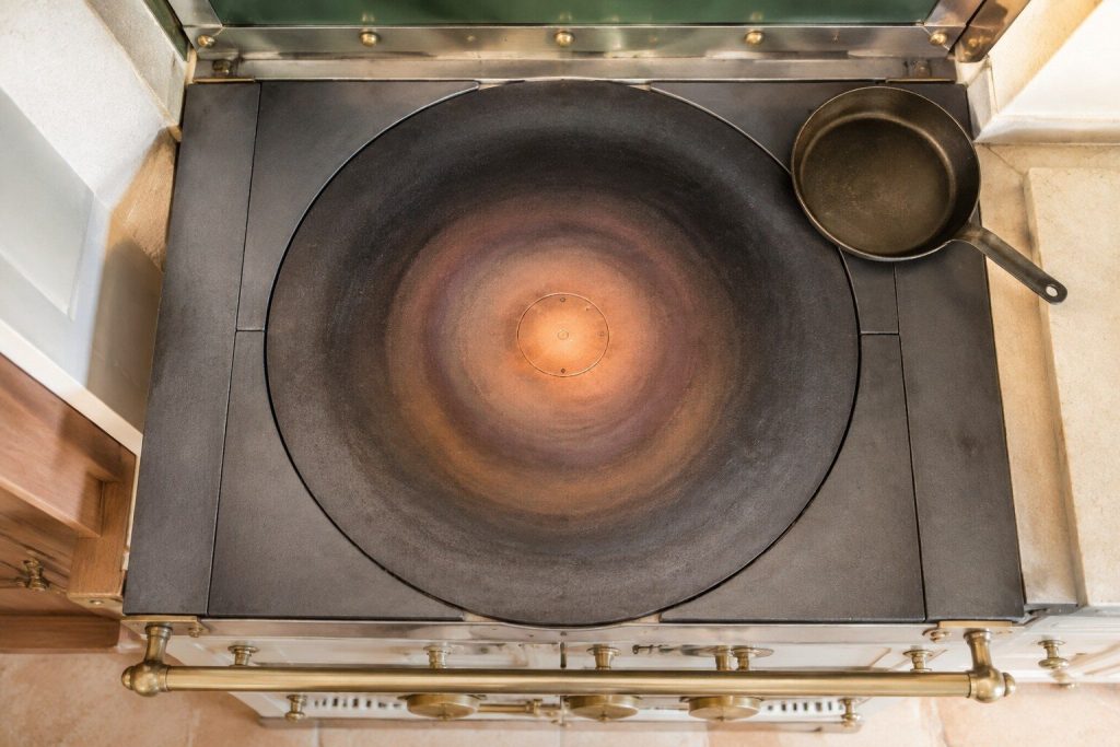 A thick round cast-iron cooking plate seen from directly above in daylight, concentric bands of discoloration spreading from a bright hot center to a dull cold rim, one small pan sitting well off to the outside edge.