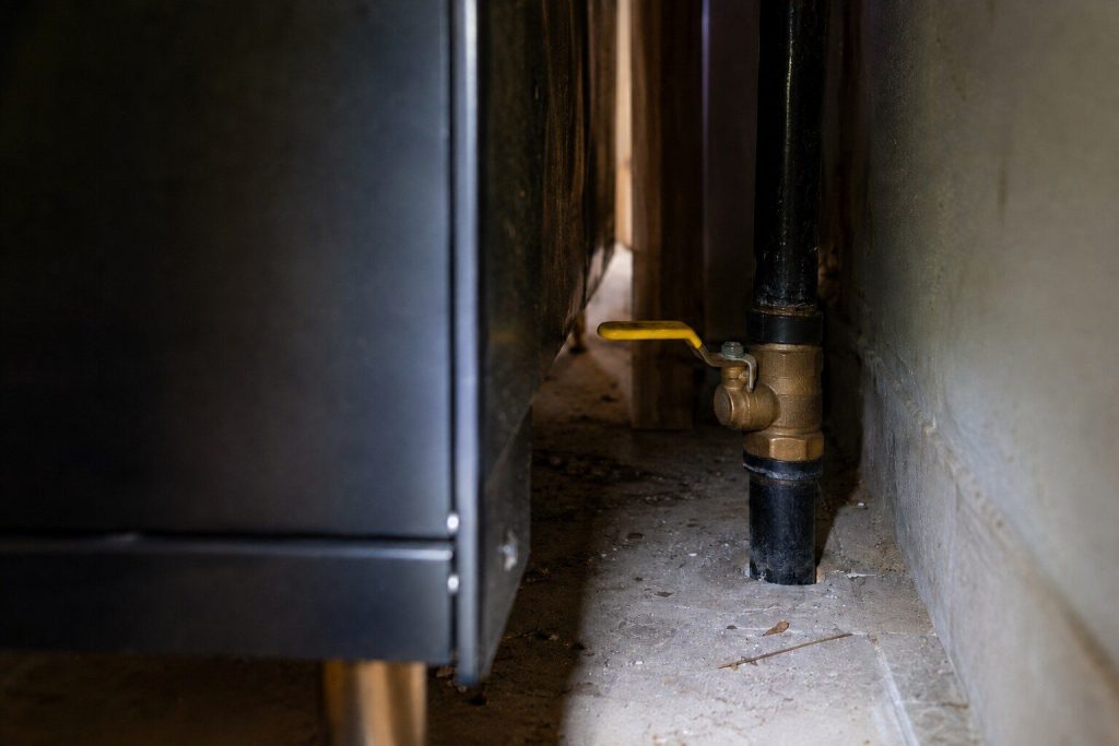 A yellow-handled quarter-turn gas valve on a black pipe in the narrow dark gap behind a range, lit by a torch beam from above, dust on the floor and the back of the appliance out of focus on the left.